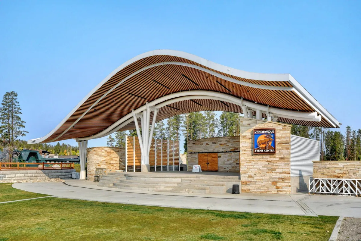 This exterior shot showcases the distinctive curved roof and stone facade of the Hideaway Park Amphitheater, with its spacious wooden deck set up for community events and performances. The outdoor setting is clearly prepared for group gatherings and concerts.