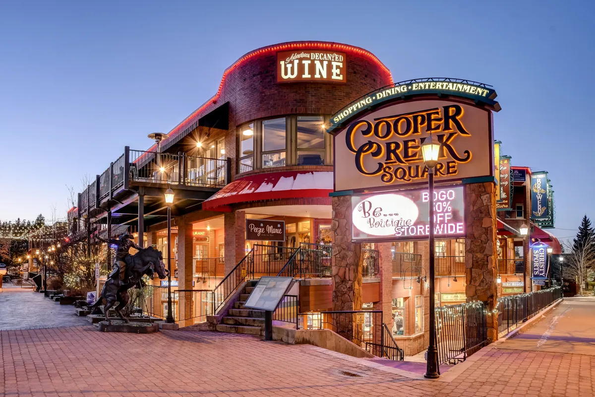 An animated exterior shot of Cooper Creek Square with visible signage for shops and wine bars, outdoor dining spaces, and a public sculpture of a horse. The setting is festive and vibrant, illustrating the dynamic urban environment just steps from the home.