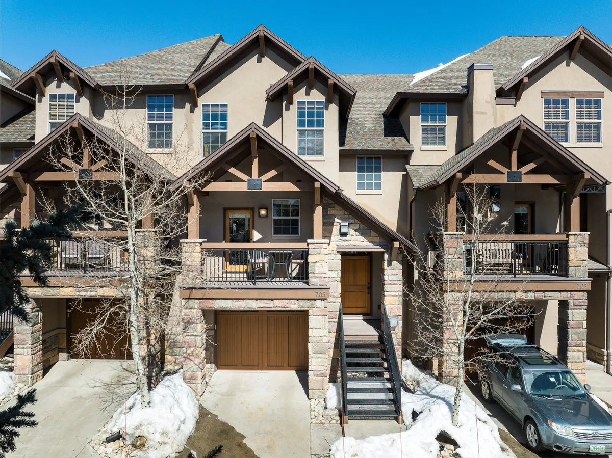 The image showcases the inviting exterior of a luxury villa nestled among aspen trees in Winter Park, Colorado. The house features distinctive wooden balconies, a striking stone facade, and welcoming front steps. Visible outdoor seating areas hint at opportunities for relaxation and enjoying the fresh mountain air. The natural setting creates a cozy, welcoming atmosphere perfect for gatherings or quiet outdoor moments.