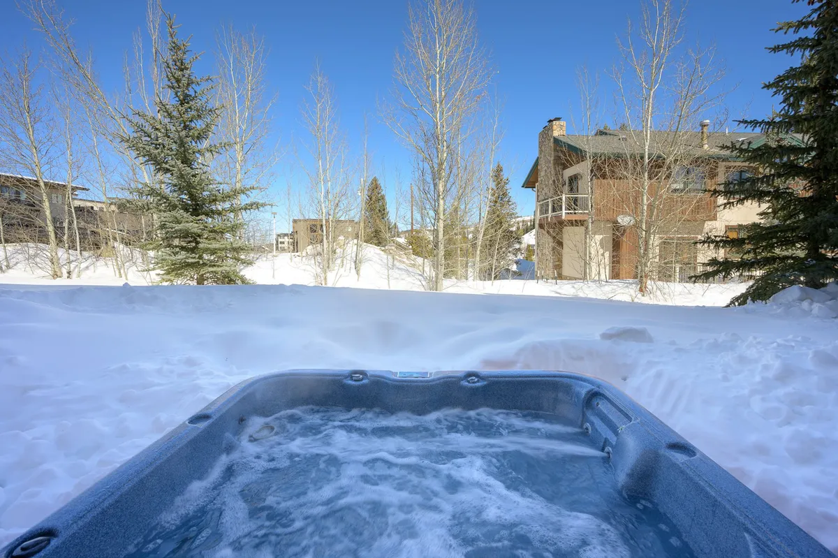 A snow-covered outdoor deck featuring a bubbling hot tub surrounded by trees and distant buildings, creating a tranquil winter retreat perfect for relaxing after a day of mountain adventures.