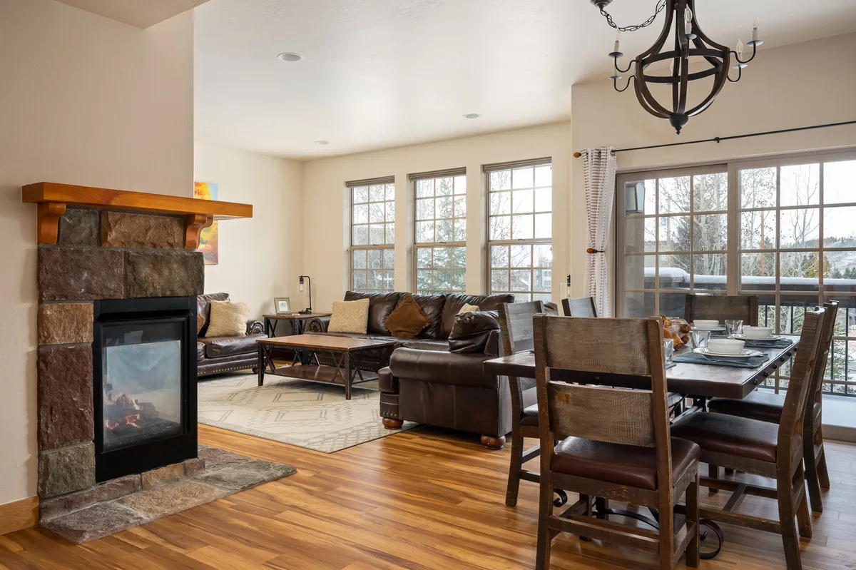 A warm and inviting open-concept living and dining area featuring a beautiful fireplace, elegant dining table, comfortable living room seating, and large windows that draw in natural light. The scene embodies the perfect blend of modern alpine design and cozy ambiance, inviting guests to gather, relax, and celebrate together.