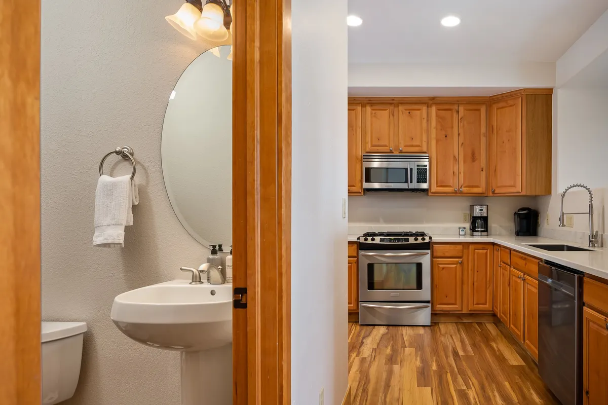 A stylish kitchen with wooden cabinetry, gleaming stainless steel appliances, and a glimpse of a mirrored bathroom sink and contemporary light fixtures. The space exudes a premium, modern feel—perfect for both family meals and sophisticated entertaining.