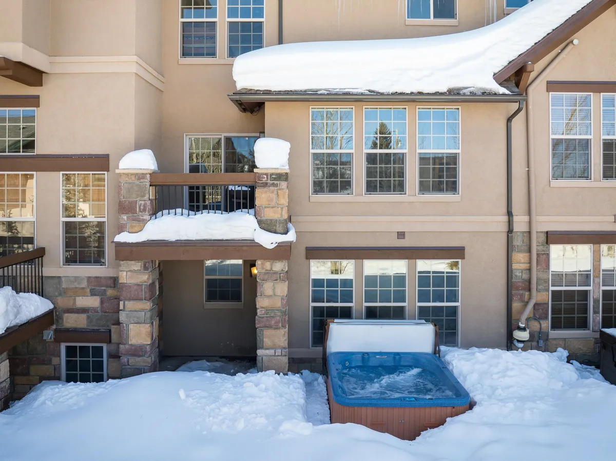 A cozy hot tub on an exterior deck with snow on the ground and a view of the home’s windows, inviting guests to relax and socialize while surrounded by Alpine beauty.