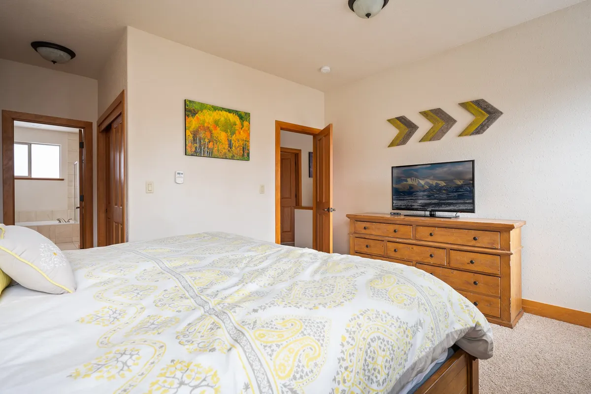 A comfortable bedroom with a patterned bedspread, wooden dresser, and a TV mounted above with artwork, creating a modern yet cozy feel. Morning light streams through nearby windows, perfect for unwinding with a show or a good book.