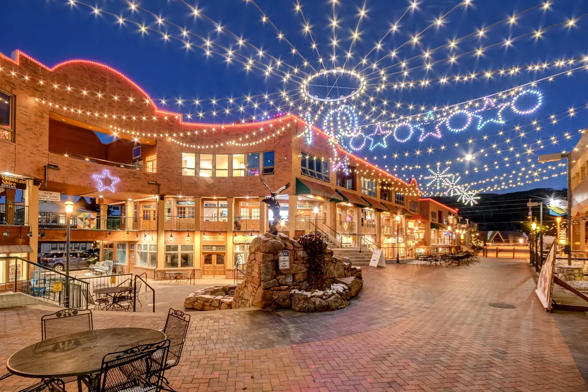 This image captures a lively outdoor plaza illuminated by string lights, with decorative stars, a stone fountain, and a welcoming seating area. The scene is bustling with people dining al fresco, shopping, and socializing beneath the glowing lights, creating a festive community atmosphere.