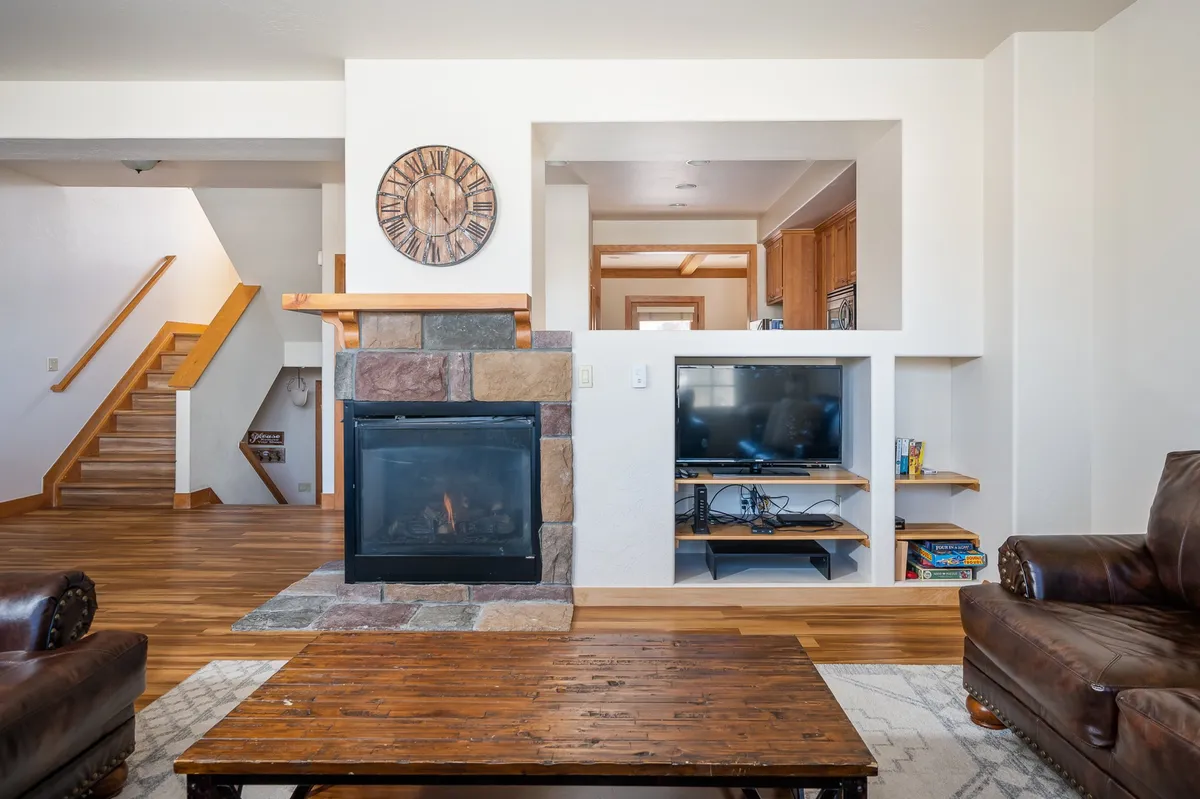A spacious, open-concept living area with a visible fireplace, leather armchairs, a wooden coffee table, a wall clock, and a seamless sightline into the kitchen. The room feels both inviting and functional, making it perfect for family gatherings and relaxed evenings.