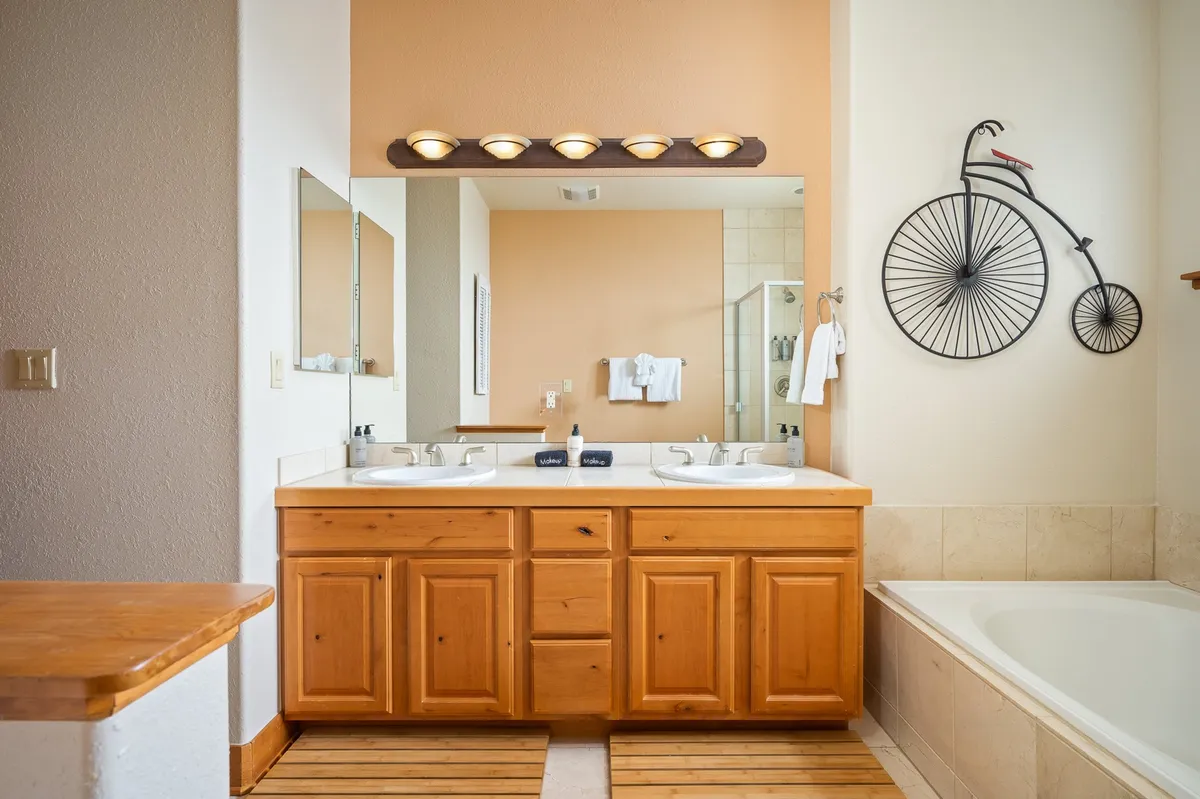 A spacious bathroom featuring a double sink vanity, framed mirrors, decorative bicycle wall art, and a soaking bathtub. The atmosphere is calm and refreshing, ideal for morning routines or relaxing baths after a busy day.